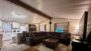 A living room with a sectional sofa, a coffee table, a rug, a TV, and a kitchen in the background. - Mountain House - Mt. Rainier Cabin - Hot Tub, Fire Pit, Pet Friendly. (Ashford)