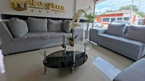 A modern hotel lobby with a grey sofa, a glass coffee table with a pineapple centerpiece, and a wall-mounted sign reading 'Regina Hotel'.