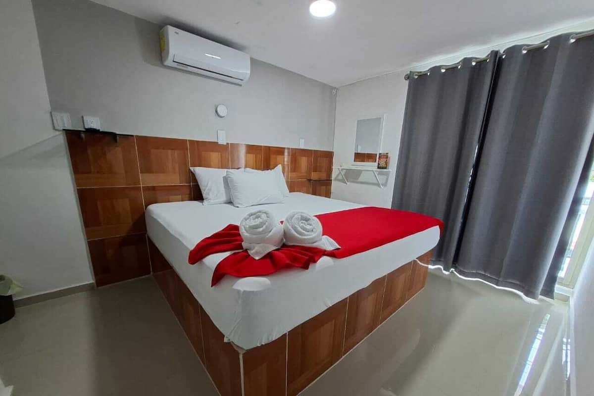 A hotel room with a bed, a red blanket, two rolled towels, a wooden headboard, an air conditioning unit, a mirror, and grey curtains.