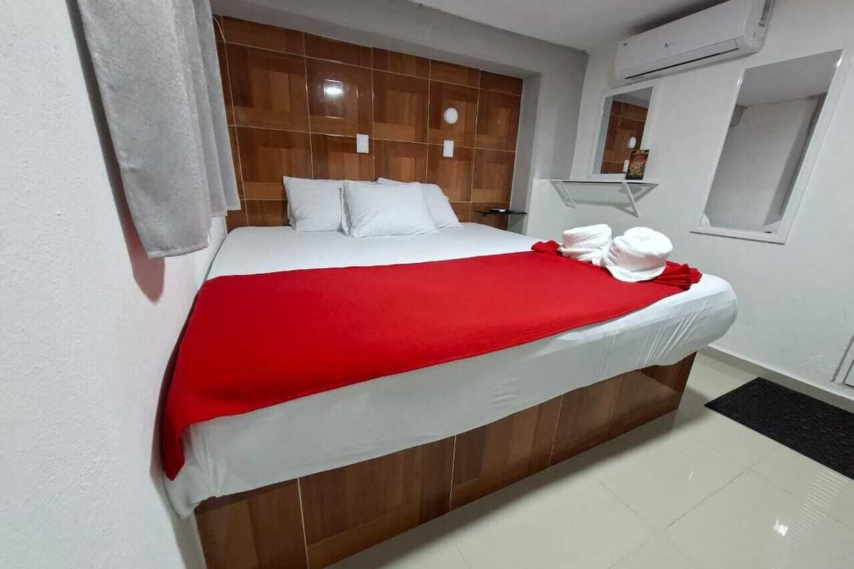 A neatly made bed with a red blanket, a wooden headboard, and a white wall.
