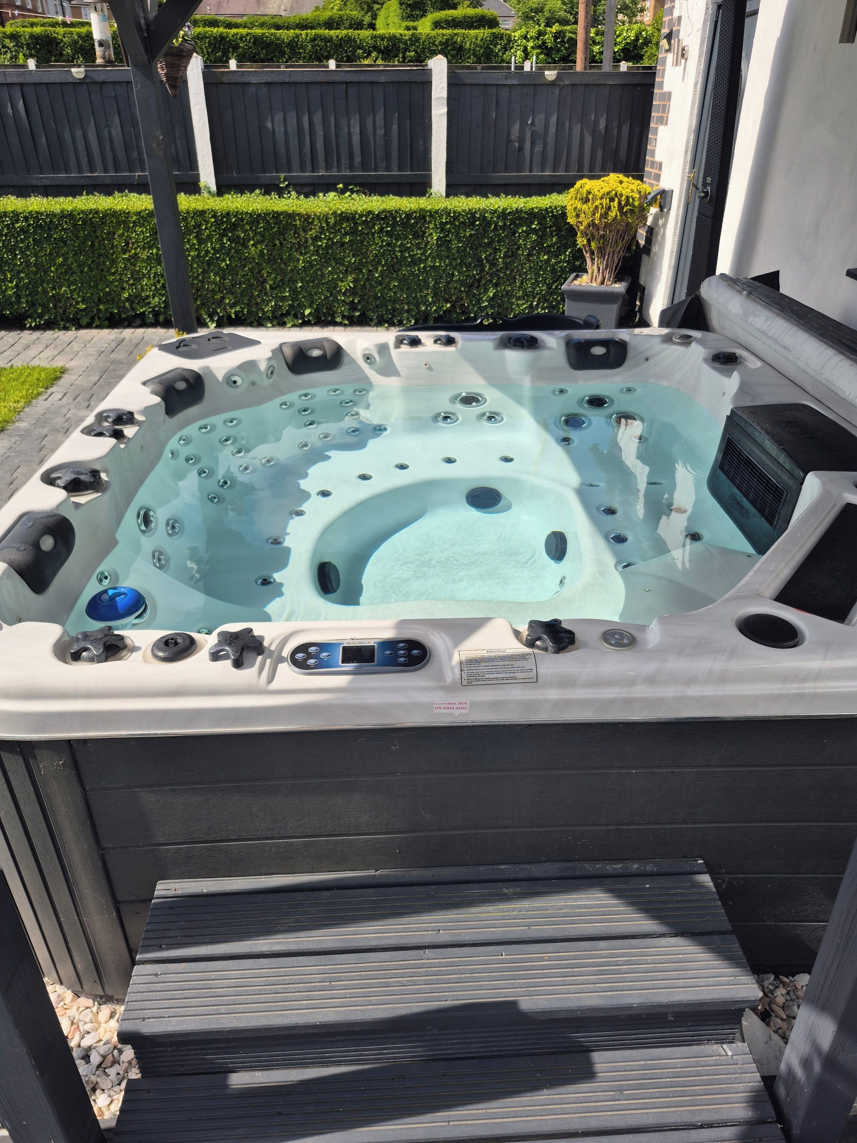 Een hottub met bedieningspaneel, omgeven door een houten terras en een haag.