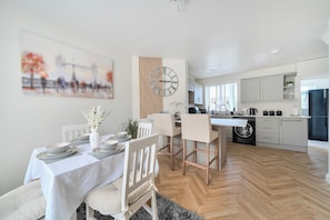 A modern kitchen with a central island, a dining area with a white table and chairs, and a large clock on the wall.
