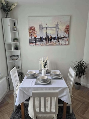 A dining area with a white table set for four, a white chair, a white cabinet with shelves, a potted plant, and a painting of a bridge. - Gym - Hot tub - Sauna - Pet - Parking - Sleeps 6 (Bagillt)