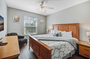 Quarto com cama, mesa de cabeceira, abajur, janela com persianas, ventilador de teto e uma obra de arte na parede.
