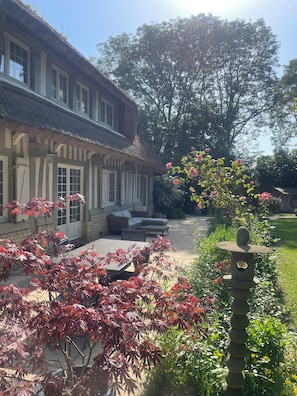 Ein traditionelles Gebäude mit einem strohgedeckten Dach, einer Terrasse mit Gartenmöbeln und einem Garten mit Pflanzen mit roten Blättern.