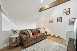 Ein gemütliches Wohnzimmer mit einem braunen Sofa, einem kleinen Beistelltisch, einer Lampe und gerahmten Bildern an der Wand.