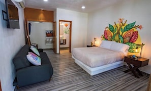 A bedroom with a bed, a sofa, a nightstand, a wall-mounted TV, and a door leading to a bathroom. - Amarxua Boutique (Acapulco)