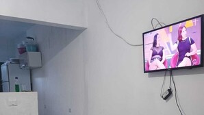Un espace de vie moderne comprenant un téléviseur à écran plat fixé au mur, un coin cuisine avec des placards et un réfrigérateur, ainsi qu’un canapé noir.