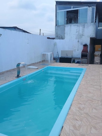 A rectangular swimming pool with a clear blue water, surrounded by a tiled patio area.. Cozy house with pool on Mongaguá beach.