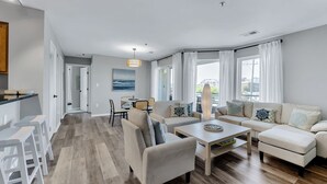 A modern living room with a sofa set, a coffee table, and a dining area with chairs. - Virginia Beach Condo With Balcony & Pool (Virginia Beach)