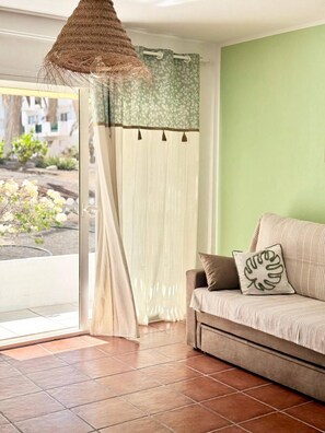 Ein Zimmer mit einer Couch, einer Hängelampe, einem Fenster mit Vorhängen und Blick ins Grüne.