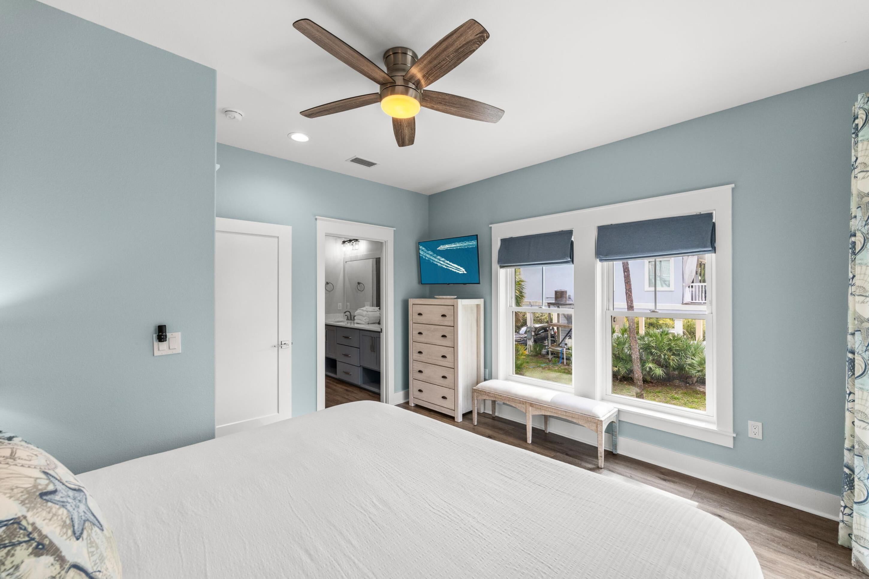 Un dormitorio con cama, ventilador de techo, ventana con vistas, cómoda y un banco.