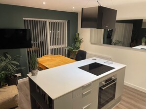 Una cocina moderna con estufa integrada, una zona de comedor con mesa de madera y un amplio ventanal con persianas.