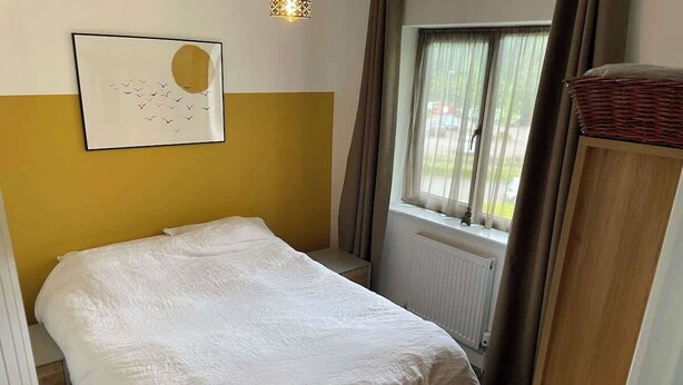 Et soveværelse med seng, et vindue med gardiner, en radiator og en vægmonteret lysarmatur.