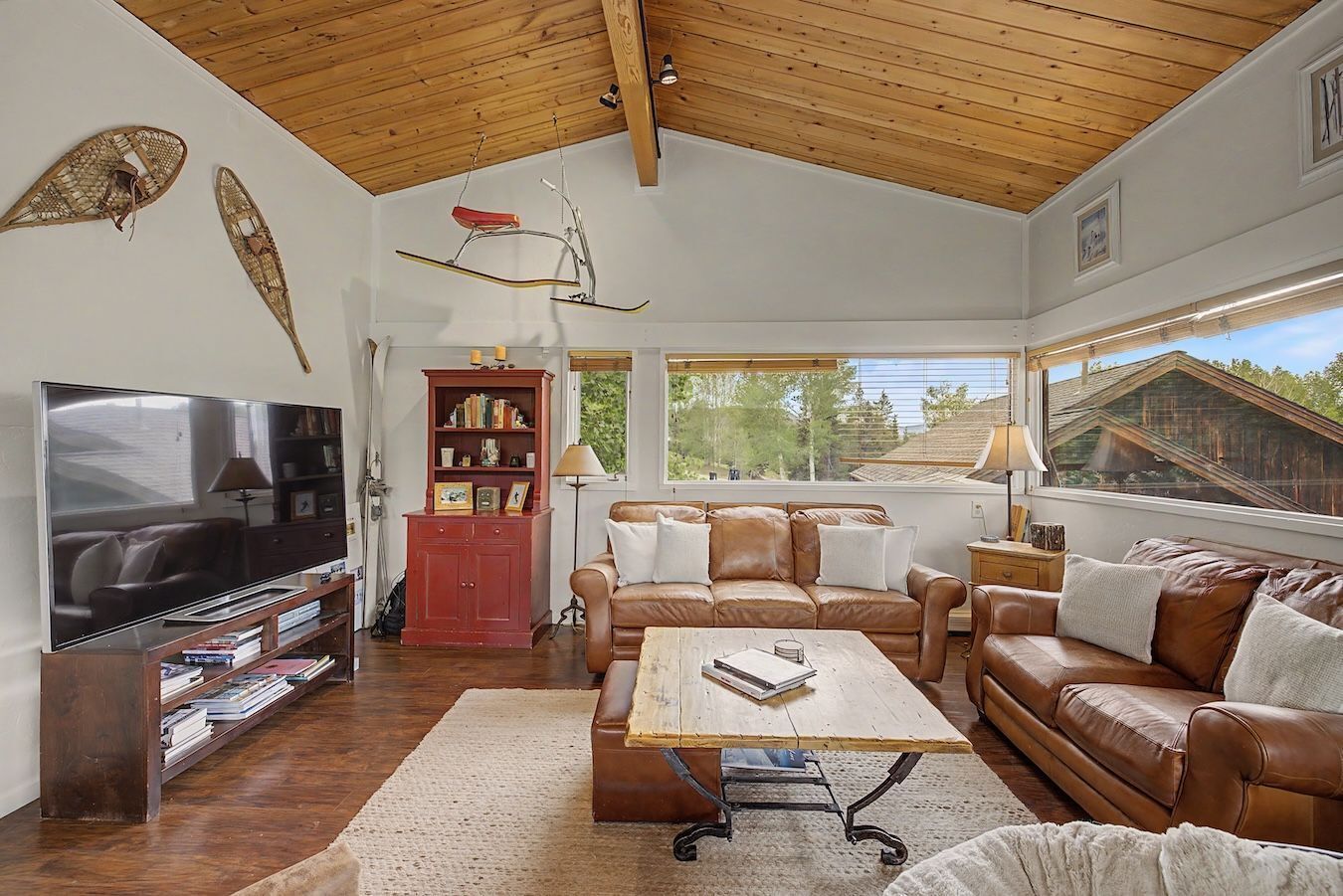 A living room with a TV, leather sofas, a wooden coffee table, a bookshelf, and a window with a view of trees.