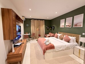 Ein modernes Schlafzimmer mit Fernseher, Holzmöbeln, einem Bett mit vielen Kissen, einem Schreibtisch mit Computer und einem kleinen Tisch mit Stuhl.