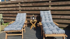 Two wooden lounge chairs with cushioned seats and backs, a small wooden table with a bottle and glasses, and a wooden fence in the background. - Carnweather (Newquay)