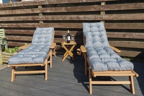 Two wooden lounge chairs with cushioned seats and backs, a small wooden table with a bottle and glasses, and a wooden fence in the background. - Carnweather (Newquay)