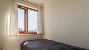 A small, single-bed room with a window and curtains, a plain wall, and a visible cityscape outside. - Carnweather (Newquay)