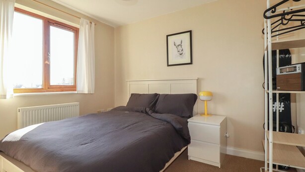 Quarto com cama, mesa de cabeceira, luminária, radiador, janela com cortinas e um suporte na parede com cabides.