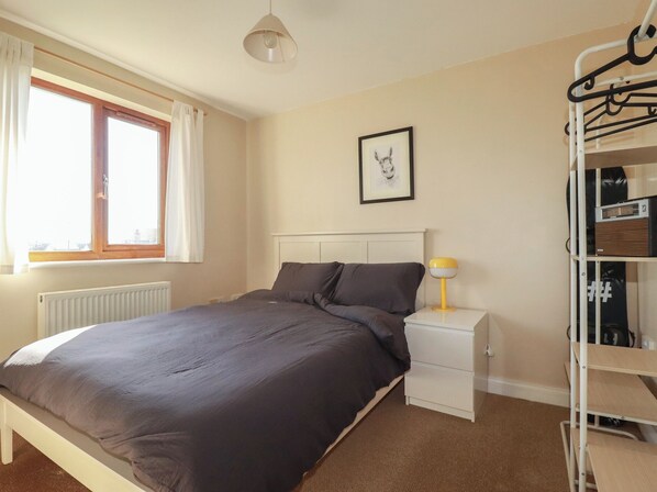 A bedroom with a bed, a nightstand, a lamp, a radiator, a window with curtains, and a wall-mounted rack with hangers. - Carnweather (Newquay)