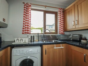 A compact kitchen with wooden cabinets, a washing machine, a sink, and a window with checkered curtains. - Carnweather (Newquay)