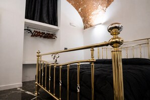 Une chambre avec un lit, une tête de lit ornée d’un cadre doré, une couvre-lit noire, un mur blanc et un rideau noir.