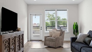 Un salon avec un téléviseur posé sur un meuble en bois, un canapé gris, un fauteuil et une grande fenêtre avec des stores.