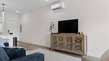 A modern living room with a flat-screen TV on a wooden cabinet, a sofa, and a dining area with a white table and chairs.