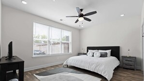 Une chambre à coucher avec un lit, un ventilateur de plafond, un téléviseur posé sur un meuble et une fenêtre avec des stores.