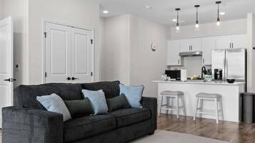 A modern living room with a dark grey sofa, white bar stools, and a kitchen area with white cabinets and stainless steel appliances.