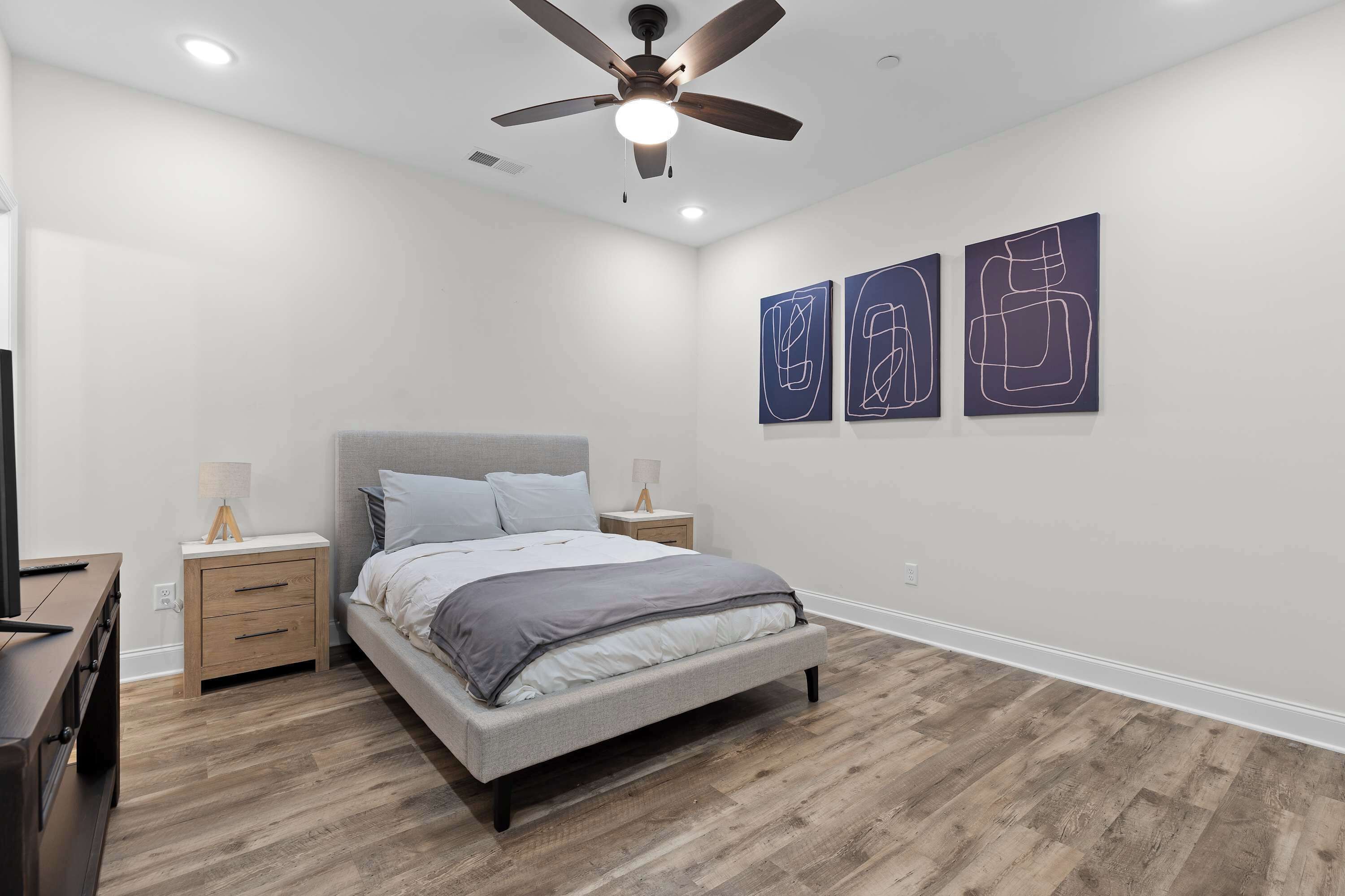 A bedroom with a bed, wooden nightstands, a ceiling fan, and abstract wall art.