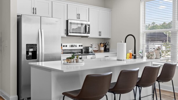 Una cocina moderna con armarios blancos, electrodomésticos de acero inoxidable, una isla central con taburetes y vistas al exterior a través de amplias ventanas.