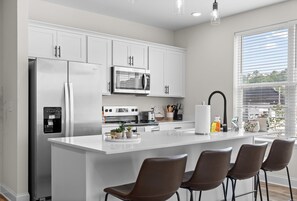 A modern kitchen with white cabinetry, stainless steel appliances, a central island with bar stools, and a view of the outdoors through large windows. - 2BR Gem in Chattanooga Southside at the Poplin (Chattanooga)