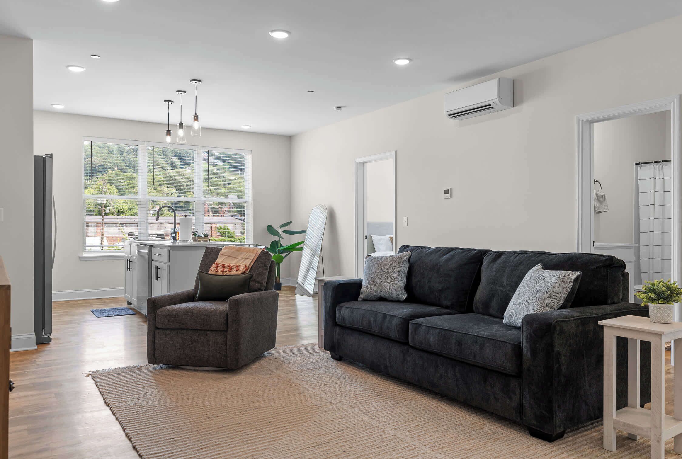 A modern living room with a dark grey sofa, a grey armchair, and a small side table. There is a large window providing natural light and a view of greenery outside.