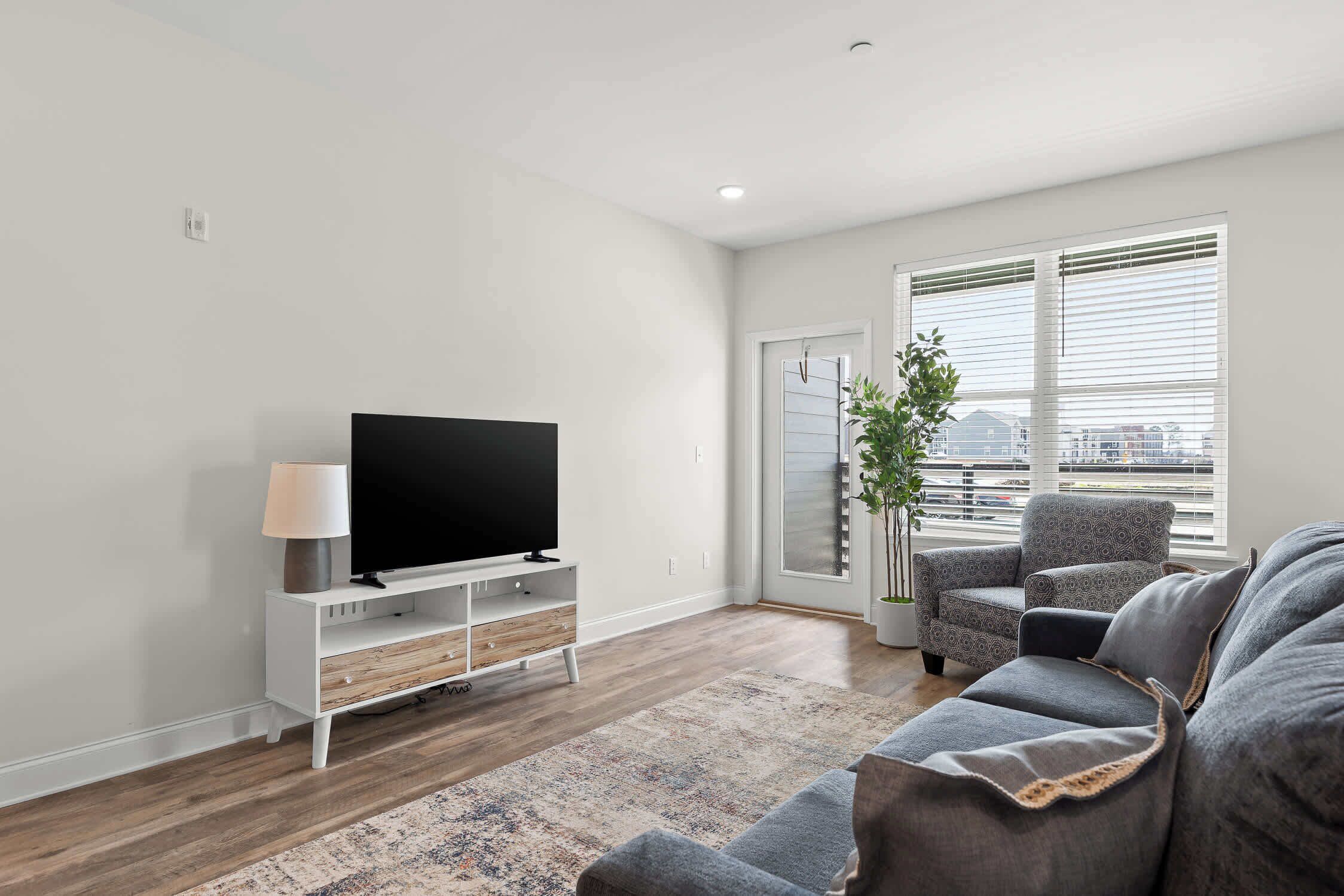 Ein modernes Wohnzimmer mit einem Flachbildfernseher auf einem weißen Schrank, einem grauen Sofa, einem gemusterten Teppich und Blick auf die Stadt durch große Fenster.