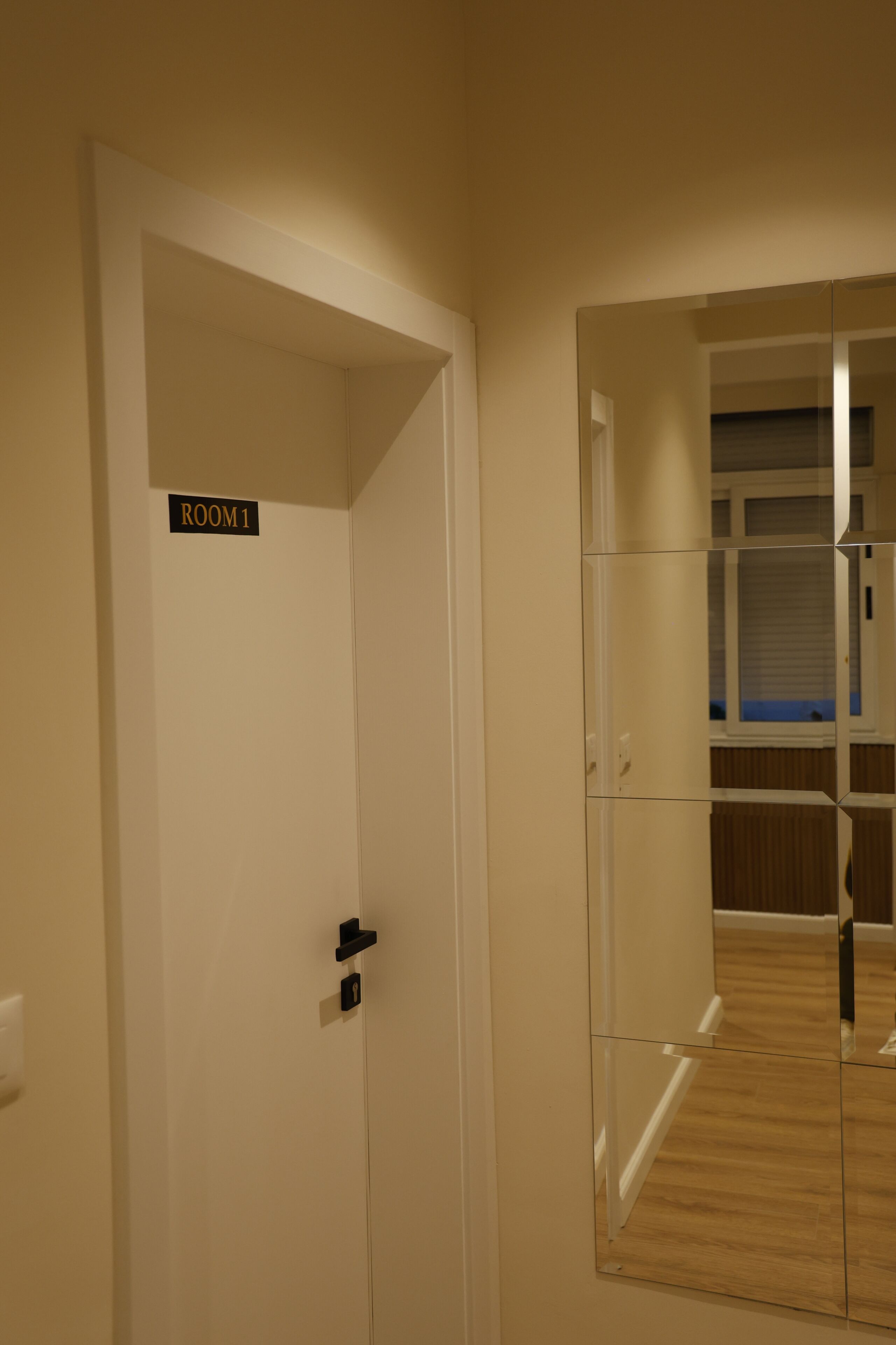 Een kamer met een deur waarop 'ROOM 1' staat, een spiegel die een hal met houten vloeren reflecteert, en een witte deuropening.