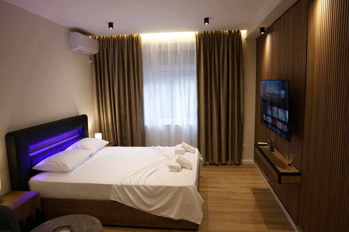 Una habitación de hotel moderna con cama, cortinas, un televisor montado y un panel de pared de madera.