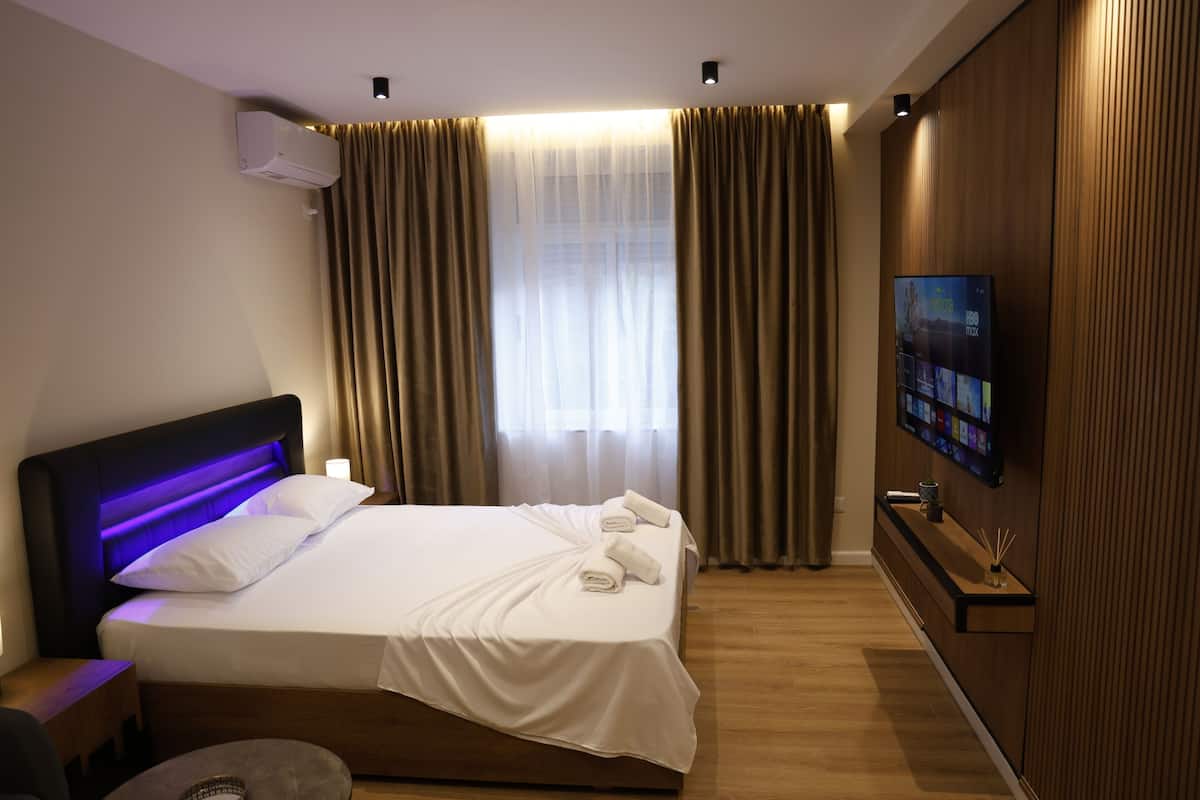 Una habitación de hotel moderna con cama, cortinas, un televisor montado y un panel de pared de madera.