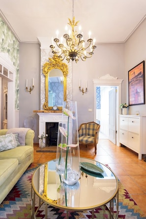 Ein Wohnzimmer mit einer Chandelüster, einem Kamin, einem Sofa, einem Glastisch und einem Sessel.