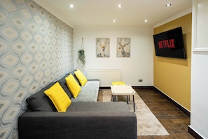 Una sala de estar moderna con un sofá gris, cojines amarillos, una pequeña mesa blanca y un televisor que muestra el logotipo de Netflix.