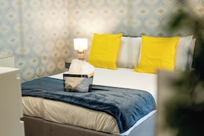 Una cama bien tendida con una colcha azul y blanca, un almohadón blanco y dos almohadones amarillo brillante.