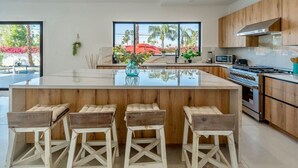 A modern kitchen with a central island, bar stools, wooden cabinets, and a view of palm trees. - Modern Villa|Palm Desert|Great location|Pool|Spa (Palm Desert)