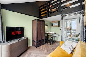 Ein Wohnzimmer mit Fernseher, der 'NETFLIX' zeigt, einem Holzkabinett, einem Esstisch mit Stühlen und einem gelben Sofa.