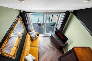 Ein Wohnzimmer mit einer Couch, einem großen Spiegel, einem Fernseher und Blick auf einen Balkon mit Gartenmöbeln.