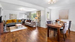 A spacious living room with hardwood flooring, a gray sofa with yellow cushions, a dining table with chairs, and a large window with curtains.
