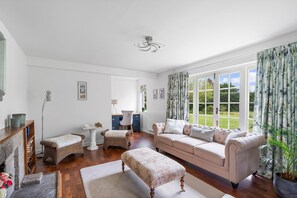 Ein Wohnzimmer mit einer Couch, Hocker, Rattanmöbeln, einem Kamin und Blick durch große Fenster.