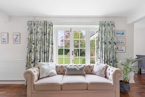 Ein beiges Sofa mit drei Kissen, ein Fenster mit blumigen Vorhängen und gerahmte Bilder an der Wand.
