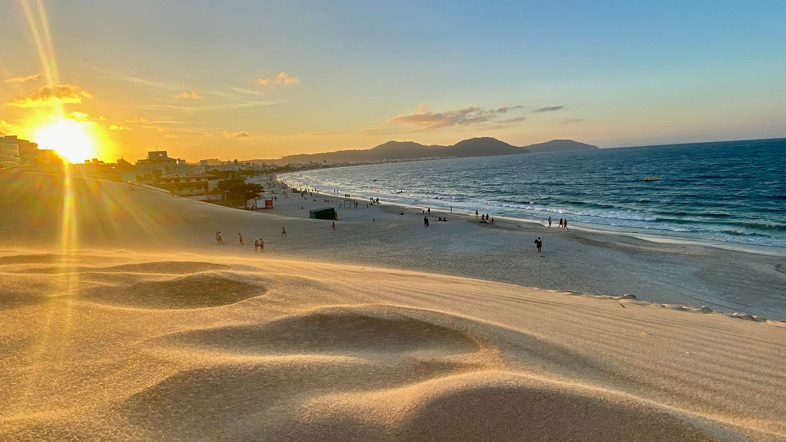 Une plage animée, un coucher de soleil et des montagnes à l’horizon.