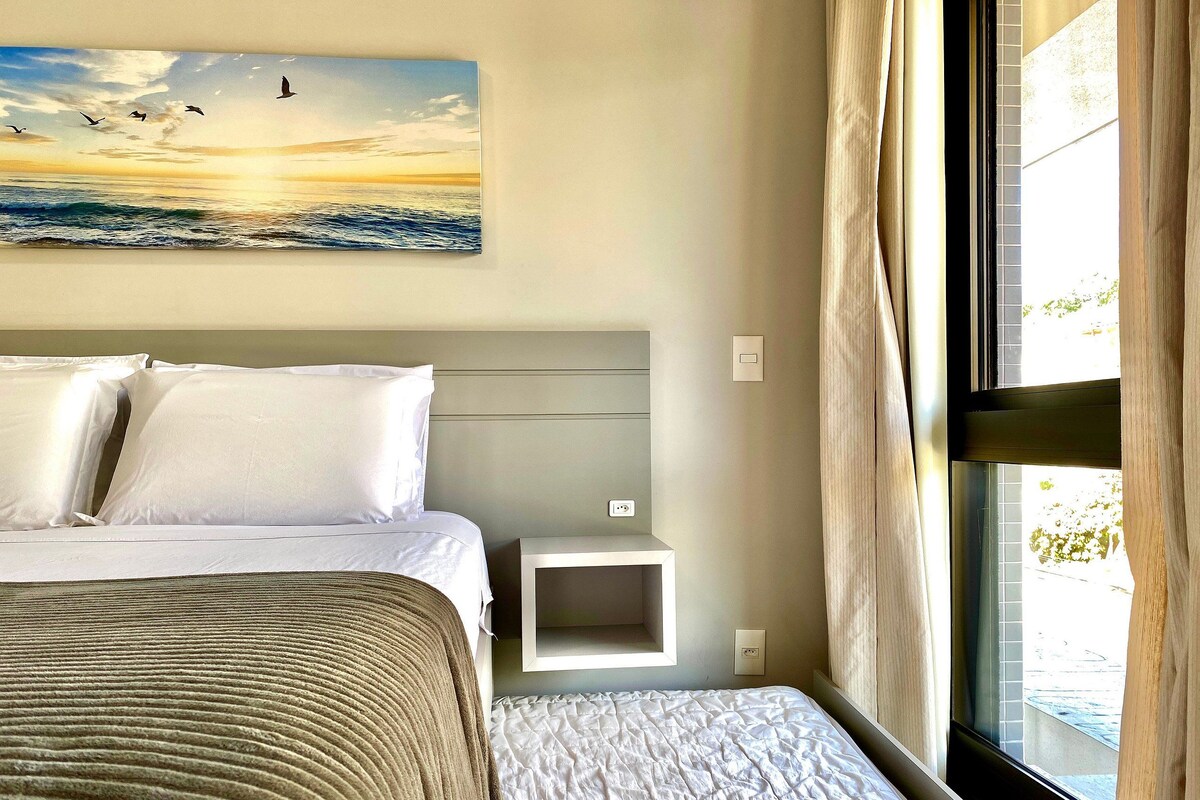 A hotel room with a bed, a bedside table, a nightstand, a painting of birds flying over water, and a window with curtains.